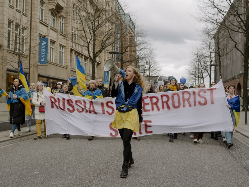 Eine Frau mit Megaphone läuft vor einem Demostrationszug mit Ukraineflagge