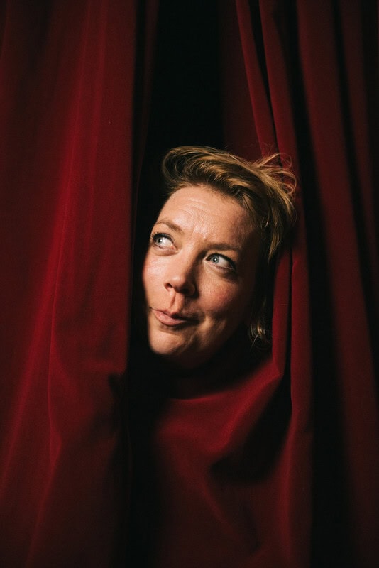 Lina Beckmann steckt den Kopf aus dem Theatervorhang und grinst