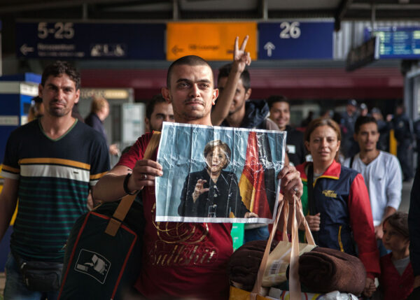 Eine Gruppe Menschen am Bahnhof hält ein Foto von Angela Merkel nach oben.