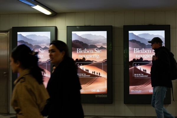 Abendliche Bahnhofswerbung für Reise-Newsletter, Menschen gehen vorbei, Stadtlandschaft.